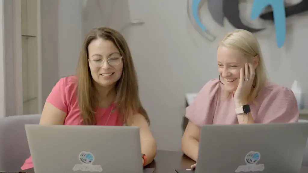 Two smiling women working on laptops at a desk in an office.