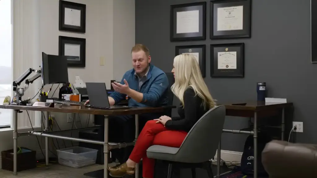 A man and a blonde woman collaborating at an office desk with framed certificates on the wall.